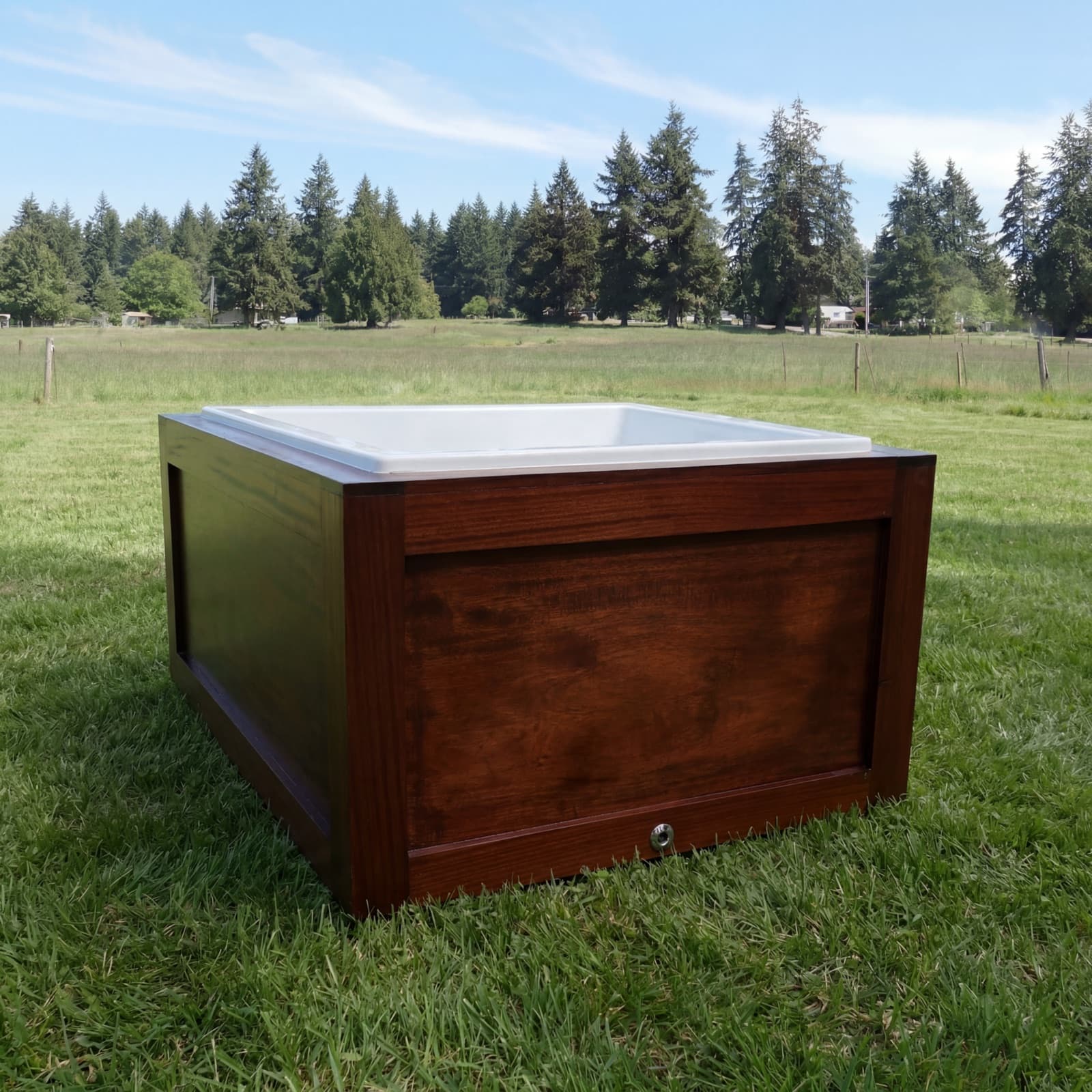 Three-quarter rear view of the mahogany cold plunge in the meadow, drain fitting low on the side panel, white basin at the brim