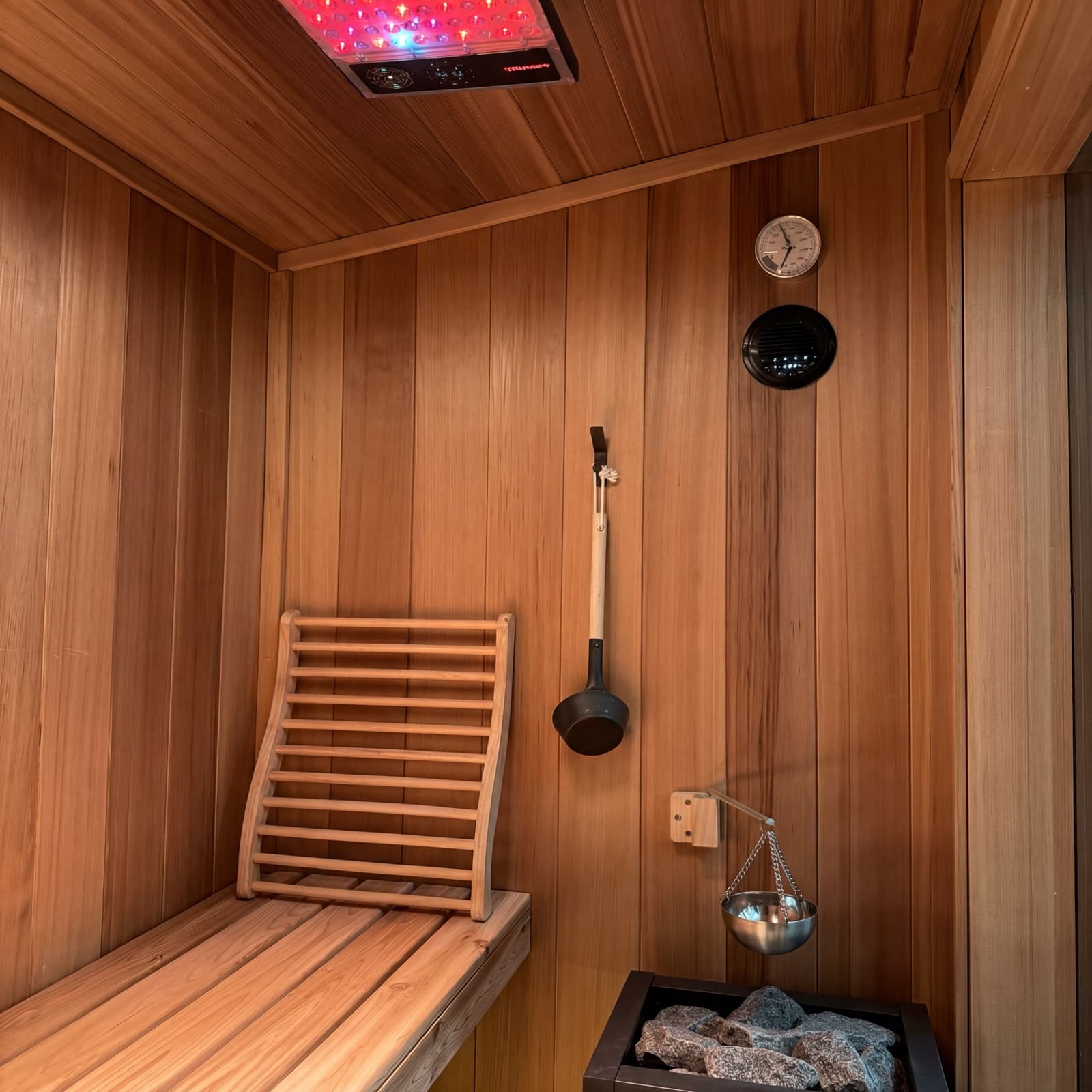 Cedar sauna interior — red light therapy panel overhead, contoured backrest on the bench, ladle and löyly bucket, stones on the heater